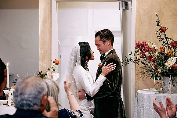 Ceremony moment as bride and groom embrace, her veil draped over a long sleeve dress in an indoor wedding ceremony with guests and aisle candles