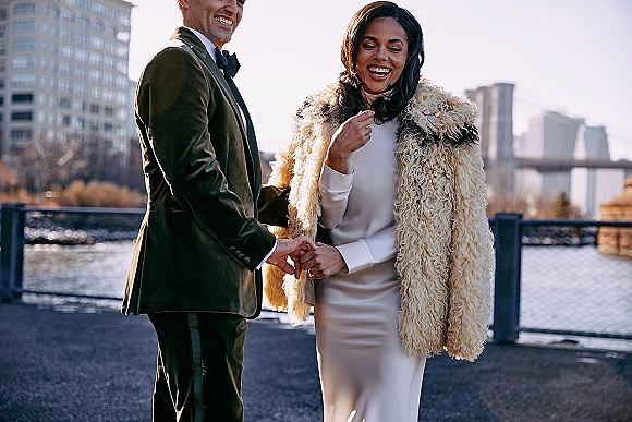 Couple portrait of bride and groom laughing, holding hands by a waterfront railing with city skyline and bridge behind them