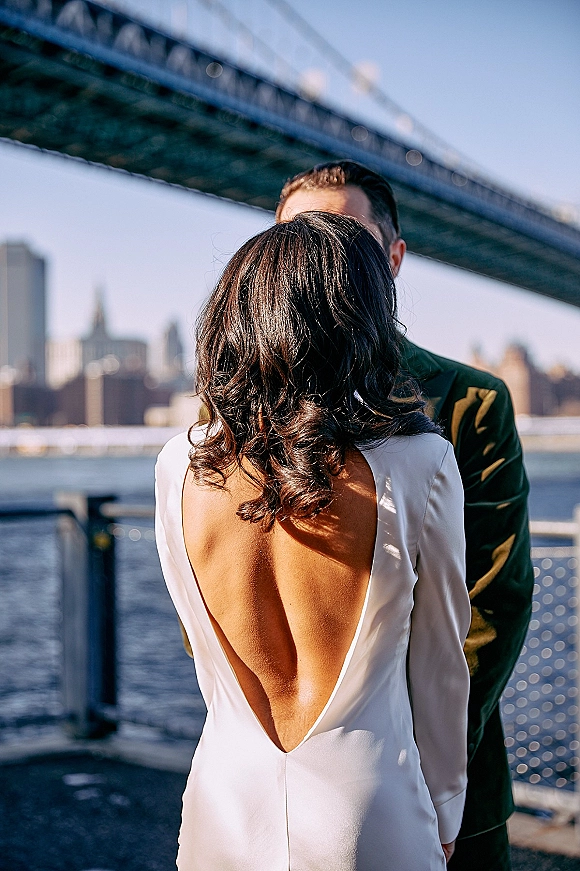 Couple portrait of bride and groom in a close embrace, bride in an open-back long-sleeve gown by a bridge with skyline beyond