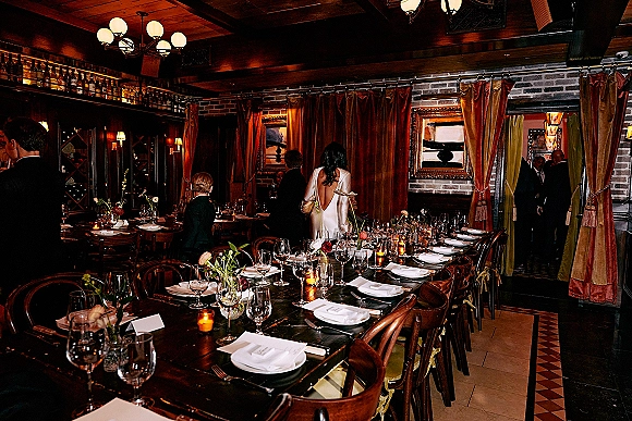 Reception tablescape with a long banquet table wedding setup, white napkins, wine glasses, votive candles and bud vases under chandeliers by a brick wall