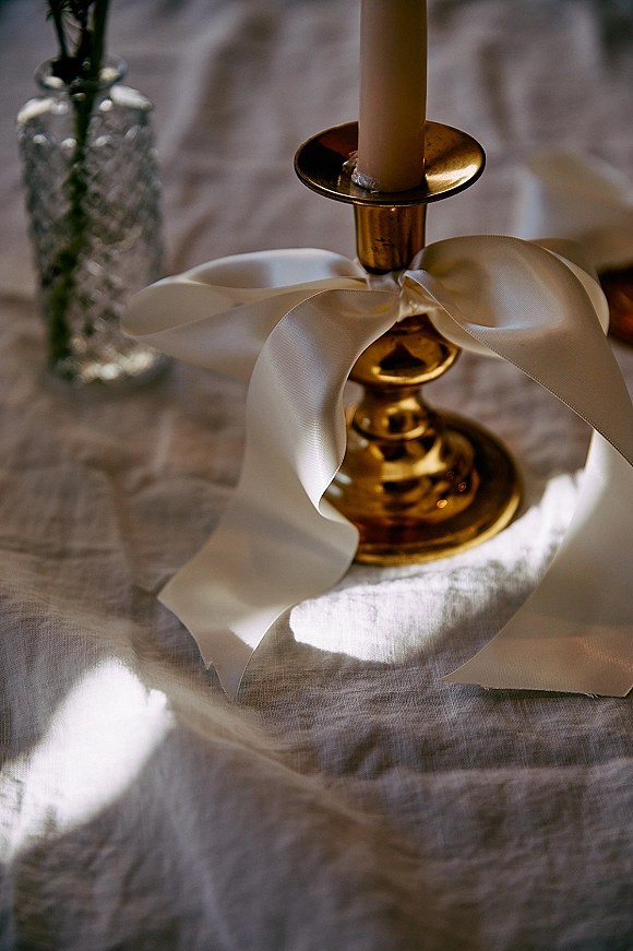 Wedding centerpiece with a taper candle on a gold candlestick, ivory satin ribbon bow, bud vase and greenery on a linen tablecloth in soft indoor light