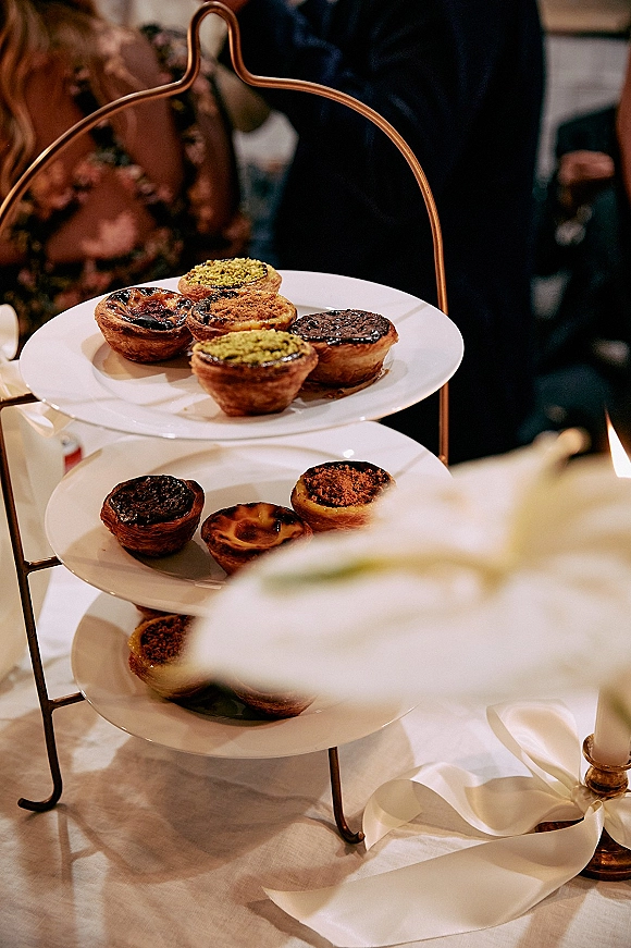 Wedding dessert table with tiered dessert stand of assorted tarts on white plates, candlelight and ribbon accents in an indoor reception space