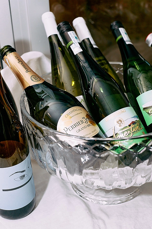 Champagne on ice in a clear glass bucket with champagne bottles and wine bottles chilling on a white tablecloth by a window
