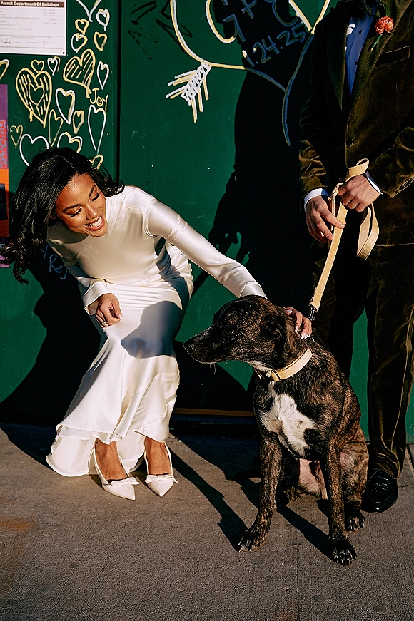 Bride with dog in a wedding dog photo, crouching in a long sleeve dress as the groom holds the leash by a green graffiti wall with heart art