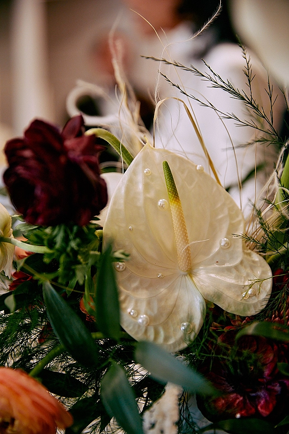 Bridal bouquet with white anthurium and burgundy flowers, lush greenery, and pearl pins, with blurred bride and groom indoors behind