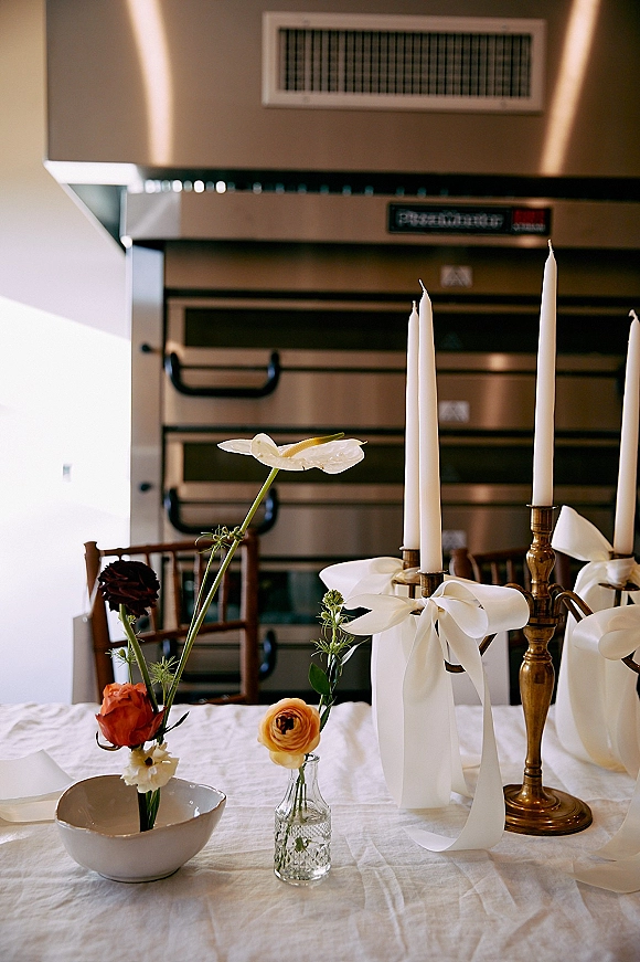 Reception tablescape with wedding table candles in brass candlesticks, white ribbon bows, bud vases, and an industrial kitchen backdrop in sunlight