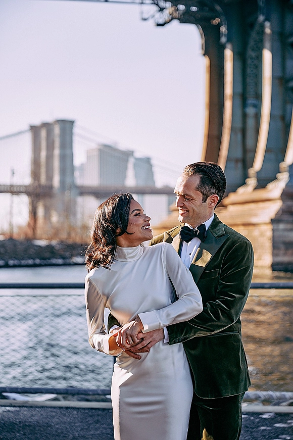 Couple portrait in a city wedding portrait style on a bridge by the river, groom in velvet tuxedo hugging bride in high neck dress, skyline behind