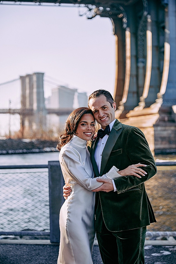 Couple portrait of bride and groom hugging by a river railing, her white satin dress and his velvet tuxedo with bow tie, city skyline behind