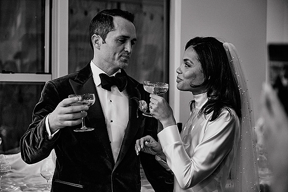Wedding toast as bride and groom clink champagne coupe glasses, her veil visible beside his tuxedo near reception windows
