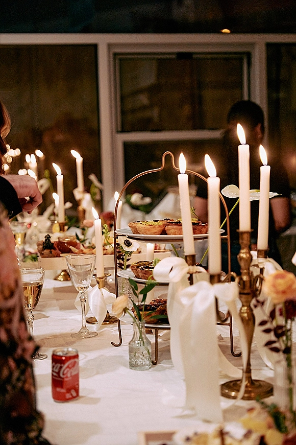 Reception tablescape with candlelit wedding table details, taper candles in brass candlesticks, ribbon bows, glassware, and guests by windows