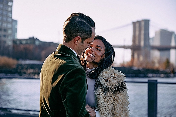 Couple portrait in a city wedding portrait, forehead touch by a waterfront railing with bridge and skyline behind, groom in velvet jacket