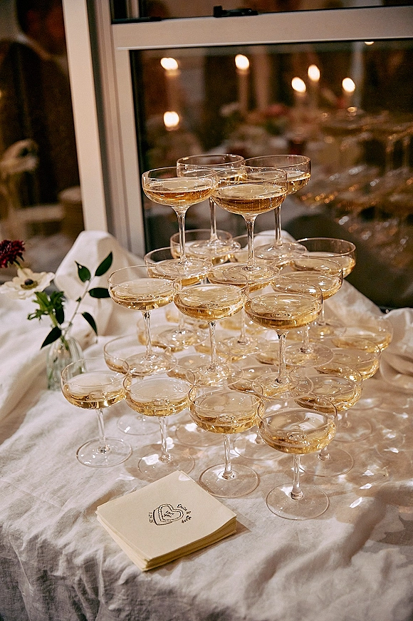 Champagne tower of coupe glasses filled with sparkling wine on a white linen table, candlelit bokeh and window in the reception background