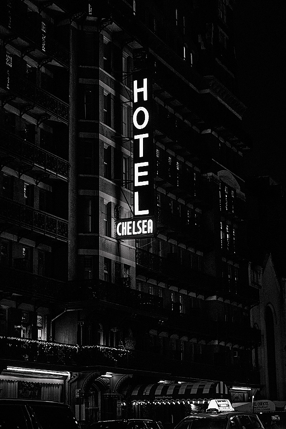 Hotel exterior with hotel neon sign glowing above a striped awning, lit windows and string lights, with cars and a taxi on the city street at night