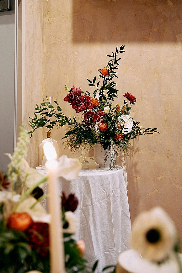 Wedding floral arrangement with orange and burgundy blooms and greenery in a ceramic vase on a linen table, taper candle, plaster wall backdrop