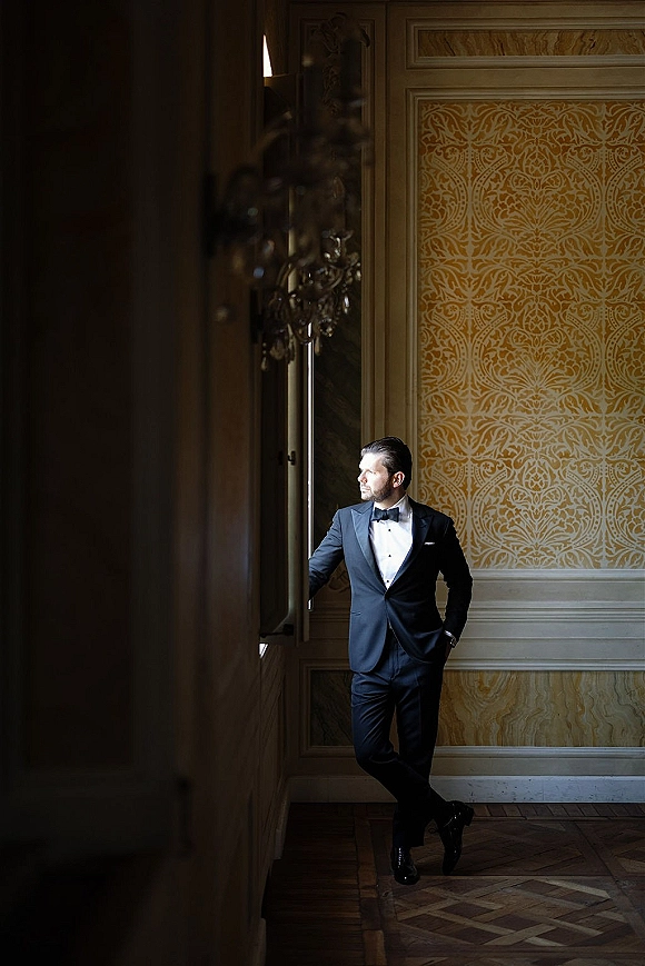 Groom portrait in black tuxedo with bow tie, standing in window light near paneled wall and patterned wallpaper in an elegant room
