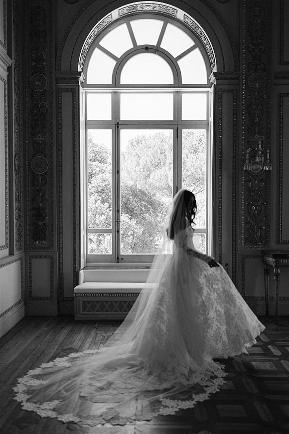 Bridal portrait in black and white of a bride by an ornate arched window, her lace wedding dress and long veil trailing on wood floors