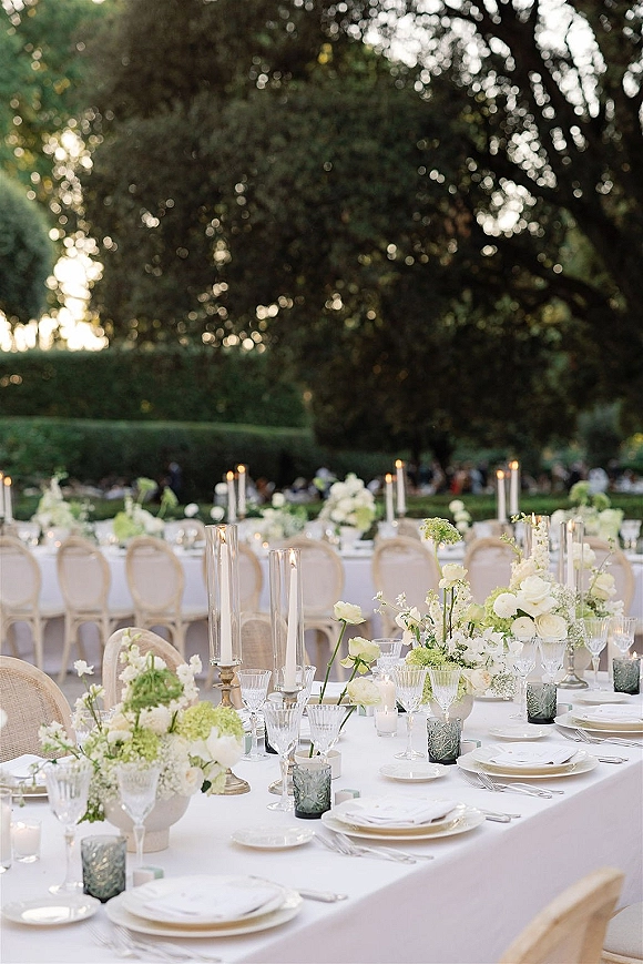 Reception tablescape with white linens, low rose and hydrangea centerpieces, taper and hurricane candles, crystal stemware on a garden lawn at dusk