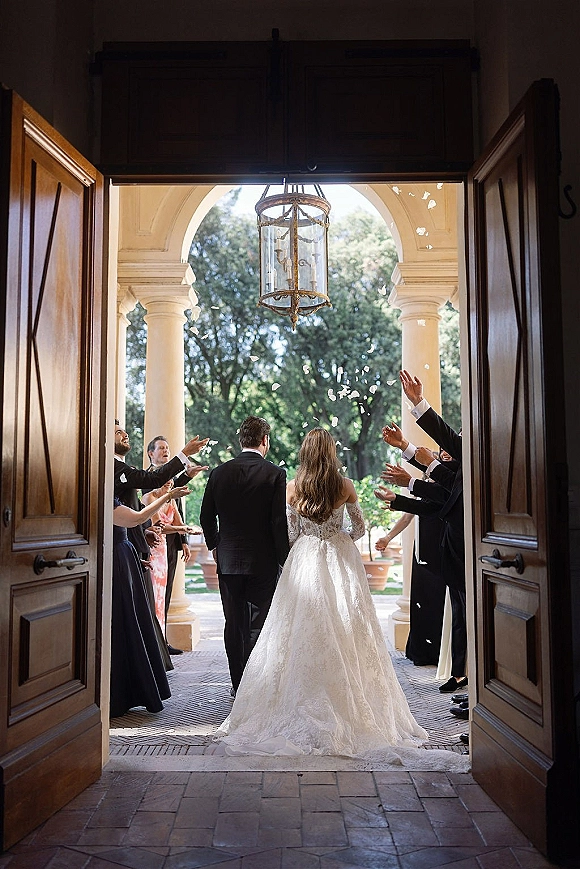 Wedding recessional with a flower petal toss as bride and groom walk away, her long train flowing through an arched doorway on stone walkway