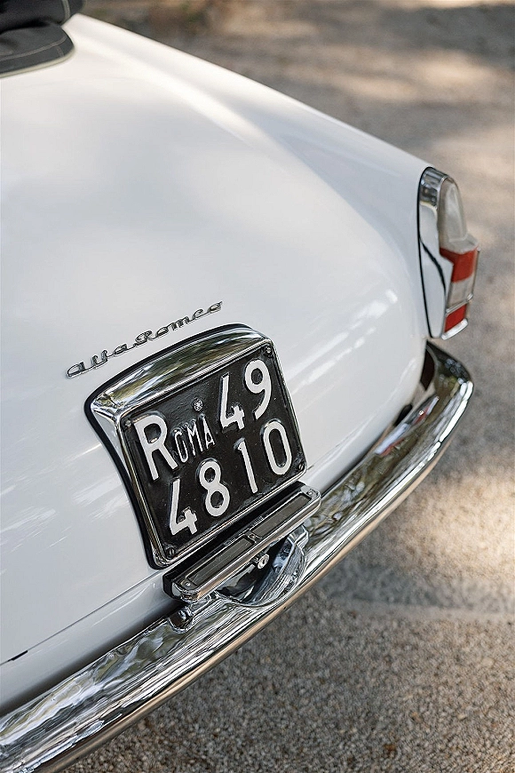 Vintage wedding car with chrome bumper and rear license plate, showing tail light and emblem on a paved road in soft shadows