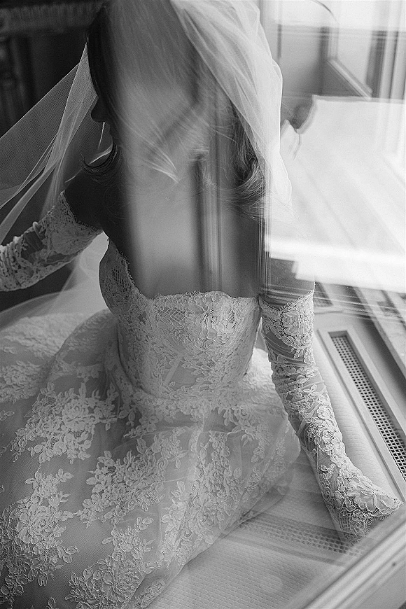 Bridal portrait in black and white of a bride by window, sitting on the floor in a long-sleeve lace dress with veil over face
