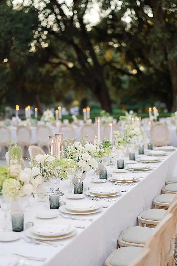 Reception tablescape with white and green floral centerpieces and taper candles on a long banquet table set outdoors in a garden setting