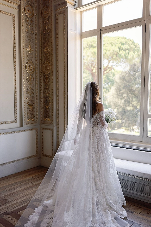 Bridal portrait of a bride by window in a lace wedding dress with a long veil, holding a white bouquet beside an ornate paneled wall
