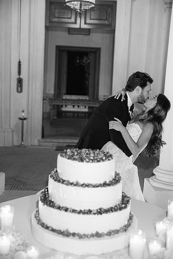 Wedding kiss as the bride and groom kissing beside a three-tier cake with candles, framed by grand interior columns and doorway backdrop