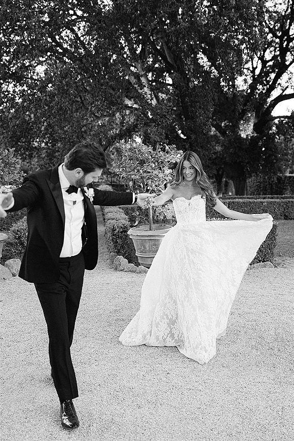 Couple portrait of bride and groom dancing as she twirls her strapless lace gown on a gravel garden path with hedges and trees