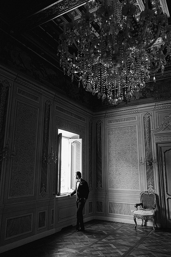 Groom portrait by window in a tuxedo with bow tie and boutonniere, standing in a luxury room with chandelier and paneled walls