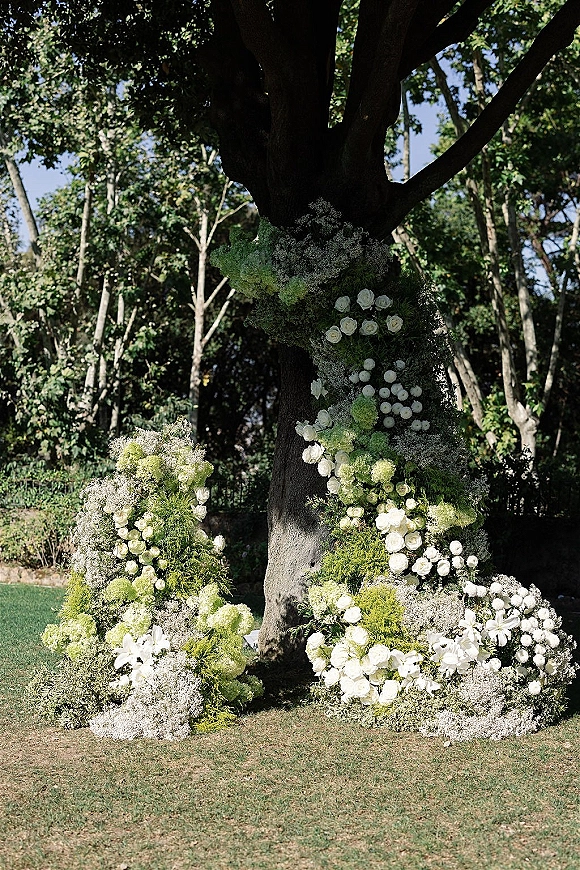 Wedding floral installation of white roses and greenery at a large tree, creating tree wedding ceremony decor on a garden lawn
