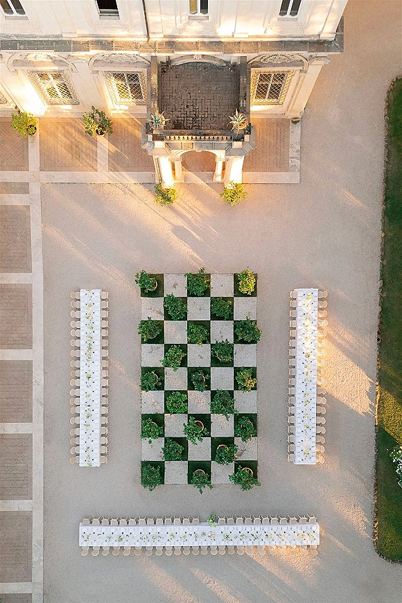 Outdoor reception layout with long white-clothed banquet tables in a U shape, set with chairs and potted greenery in a stone courtyard.