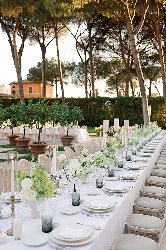 Reception tablescape with long banquet table wedding decor, white florals and hurricane candles, set on a garden lawn by a villa