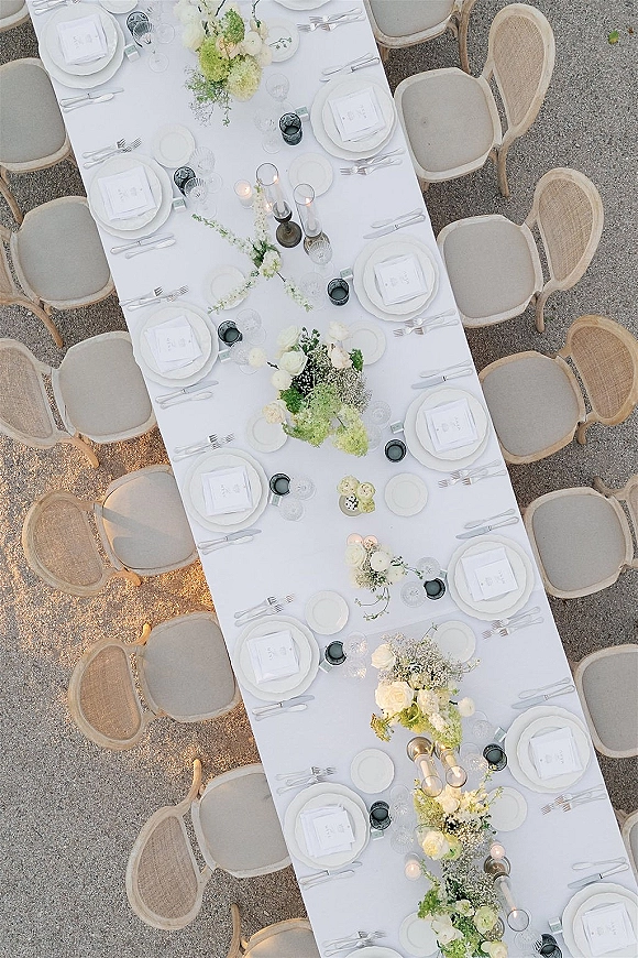Reception tablescape on a long wedding table with white linens, teal votives, taper candles, and greenery floral runner on gravel ground