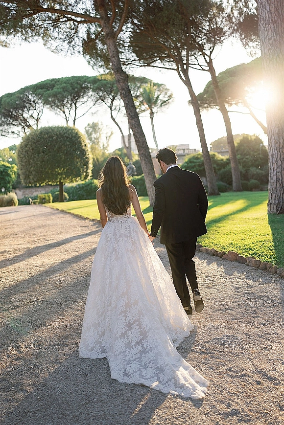 Newlywed couple walking hand in hand down a garden path, bride’s lace wedding dress train flowing beside groom in a suit in sunlight