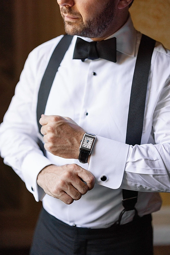 Groom getting ready, adjusting cufflinks on a white dress shirt with black bow tie and suspenders against a brown indoor wall backdrop