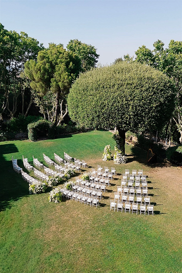 Outdoor ceremony setup with garden wedding ceremony seating in a curved aisle, white chairs and floral arrangements on a sunny lawn