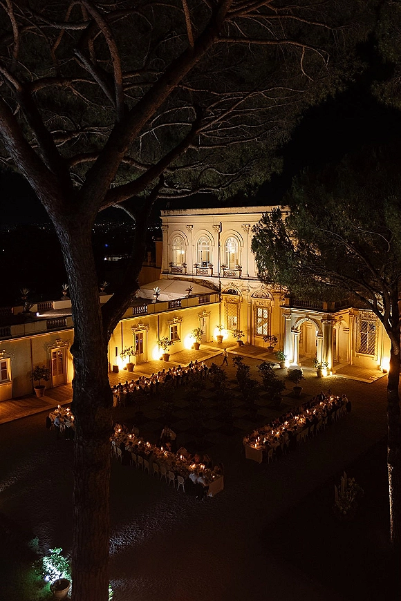 Outdoor reception setup with long banquet tables lined in candles and white linens, warm uplighting in a villa courtyard under trees at night