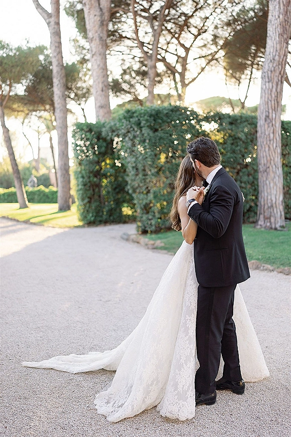 Wedding couple portrait of bride and groom embrace on a gravel garden path, her lace dress train trailing as sunlight filters through trees