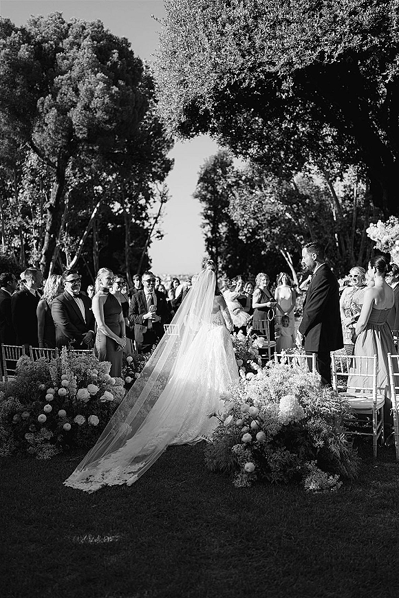 Wedding ceremony moment as the bride walks down the outdoor wedding ceremony aisle with a cathedral veil, guests standing under trees