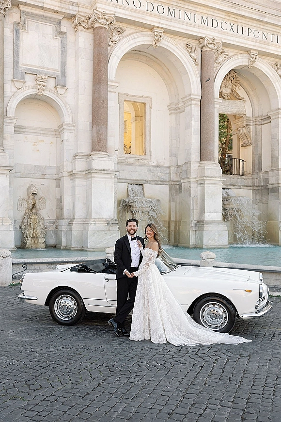 Couple portrait of bride and groom with car, holding hands beside a vintage convertible by a stone fountain and arched facade