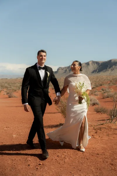 Couple portrait of bride and groom holding hands, her calla lily bouquet and veil flowing in red sand desert with mountains behind