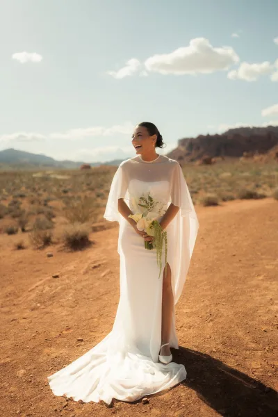 Bridal portrait of a laughing bride holding a calla lily bouquet, wearing a sheer cape veil on a red dirt road in the desert mountains