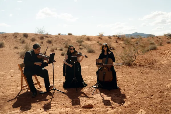 Wedding musicians play violin and cello beside music stands in black attire, set against sand dunes and distant mountains under blue sky