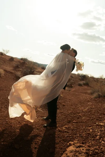 Wedding kiss portrait of bride and groom kissing in a dip, veil blowing beside a white bouquet against a rocky desert sky