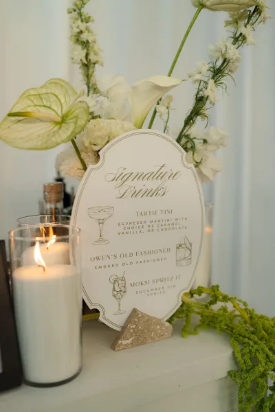 Signature drink sign with wedding signature cocktails listed in script, framed by calla lilies and white florals with candlelight on a tabletop