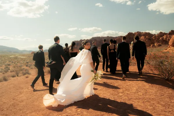 Wedding party portrait with bride looking back, long veil and calla lily bouquet, walking a desert dirt road with red rocks behind