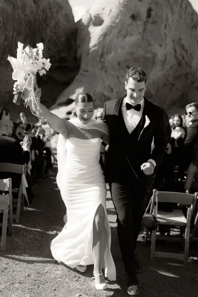 Wedding recessional as bride and groom walk the aisle, bride raising a bouquet with veil and earrings, rocky backdrop and guests cheering