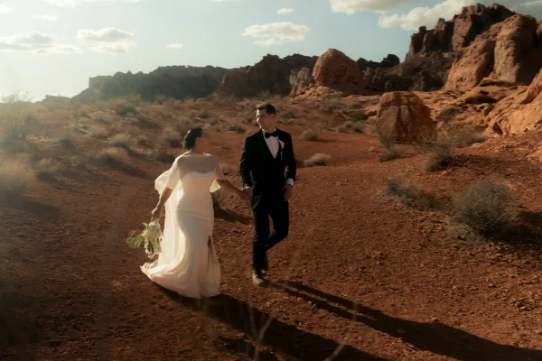 Couple portrait in a desert wedding portrait, bride in veil holding a minimal bouquet with groom in black tux, red rocks behind