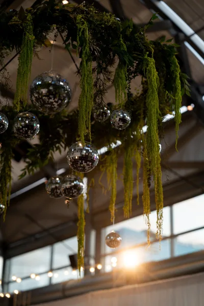 Hanging wedding installation with disco ball wedding decor, greenery and string lights suspended from metal beams in an industrial loft venue