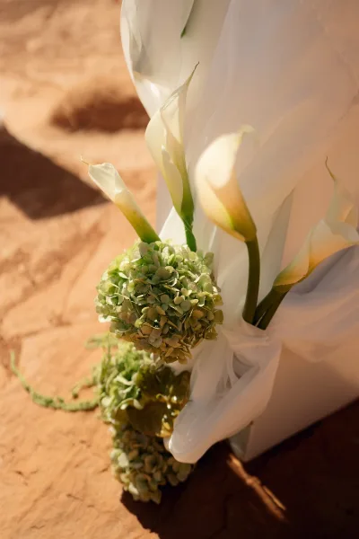 Wedding bouquet of white calla lilies and green hydrangea tied with a white ribbon, lit by warm sunlight against sandstone rocks